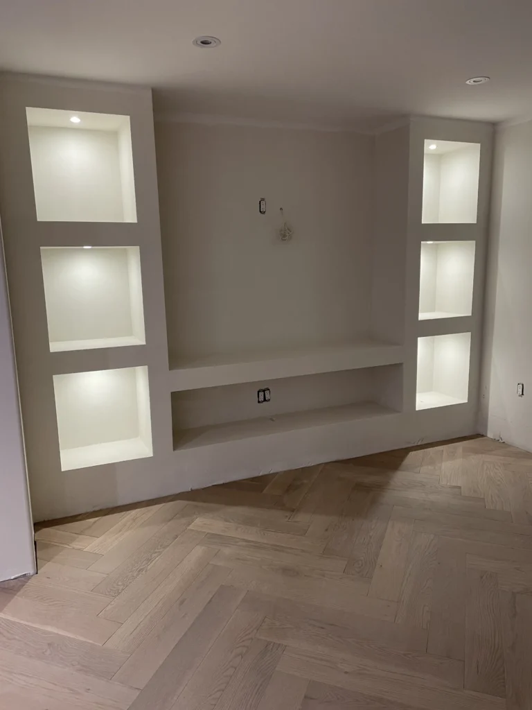 Built-in wall shelves with recessed lighting in a room with herringbone-patterned wooden flooring. 